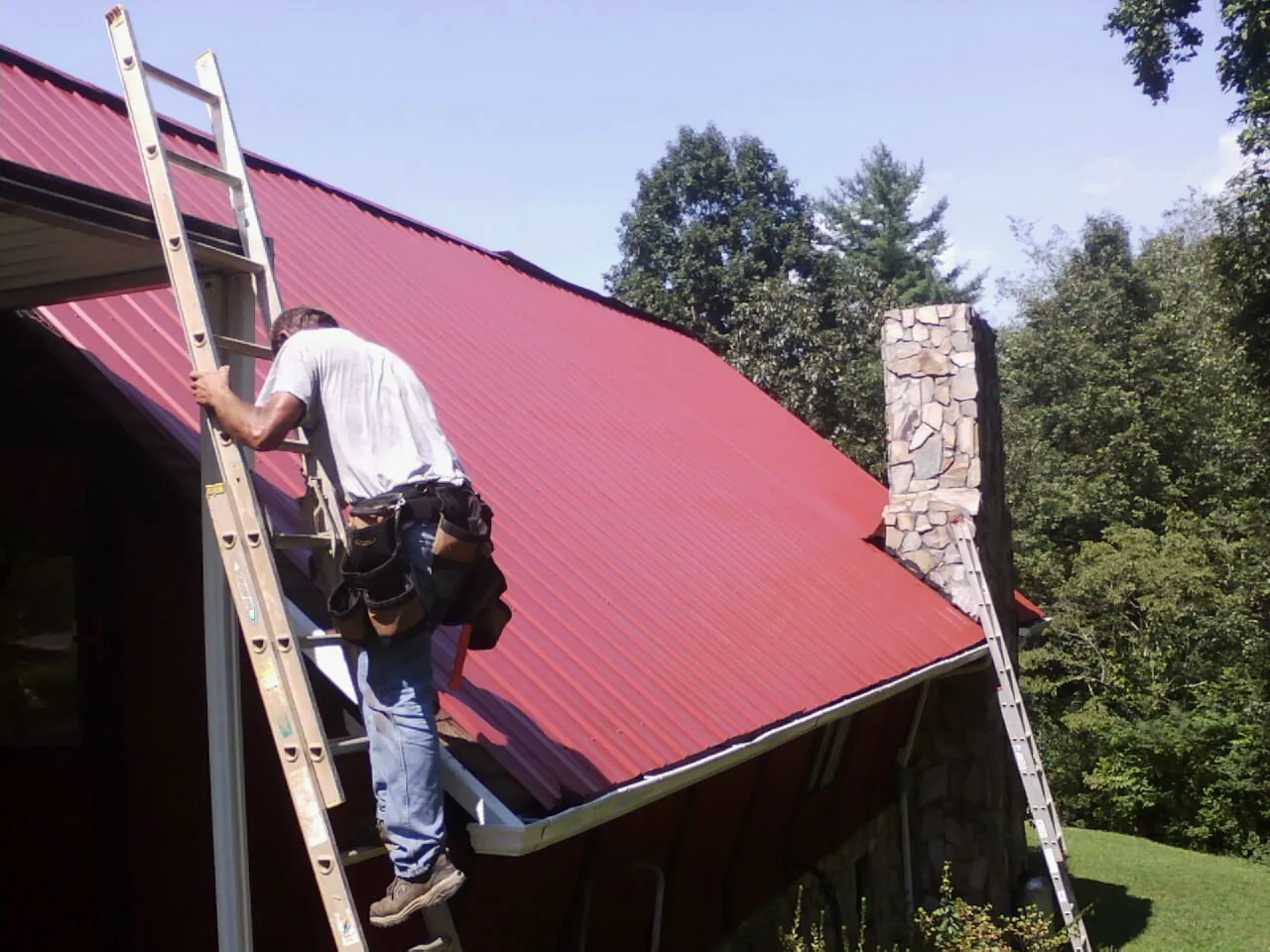 Roofer on a ladder beside a red metal roof and stone chimney — Soffit & Fascia Repair by Roofing Brothers in Briggs